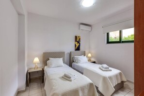 Une chambre d’hôtel avec deux lits, chacun recouvert d’une courtepointe blanche et surmonté d’une serviette pliée. Un tableau est accroché au mur au-dessus des lits, il y a une fenêtre donnant sur la verdure, et un appareil de climatisation est fixé au mur.