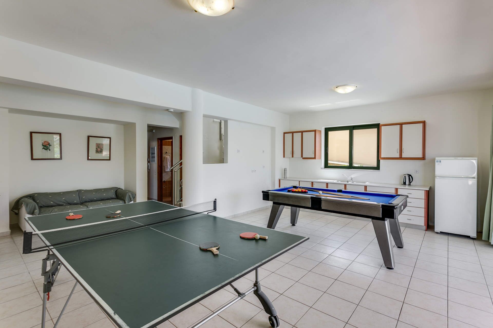 A room with a ping pong table, a pool table, a sofa, and a kitchenette.