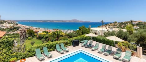 En swimmingpool med solstolar och parasoller, med utsikt över en kuststad och havet.