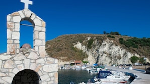 Une structure en pierre surmontée d’une croix, un arc en pierre, et une vue sur des bateaux et un paysage vallonné en arrière-plan.