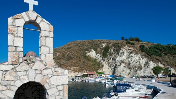 Une structure en pierre surmontée d’une croix, un arc en pierre, et une vue sur des bateaux et un paysage vallonné en arrière-plan.