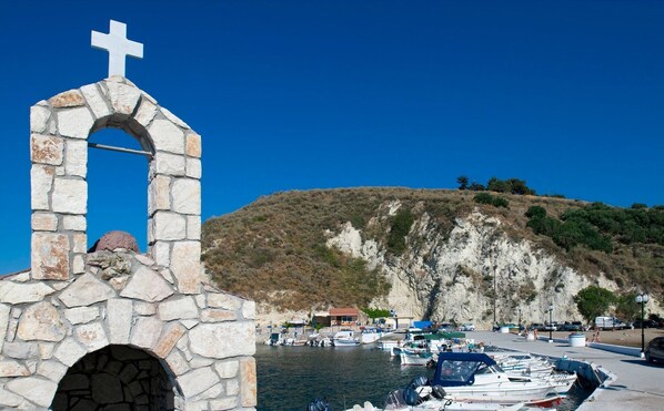 A stone structure with a cross on top, a stone archway, and a view of boats and a hilly landscape in the background.