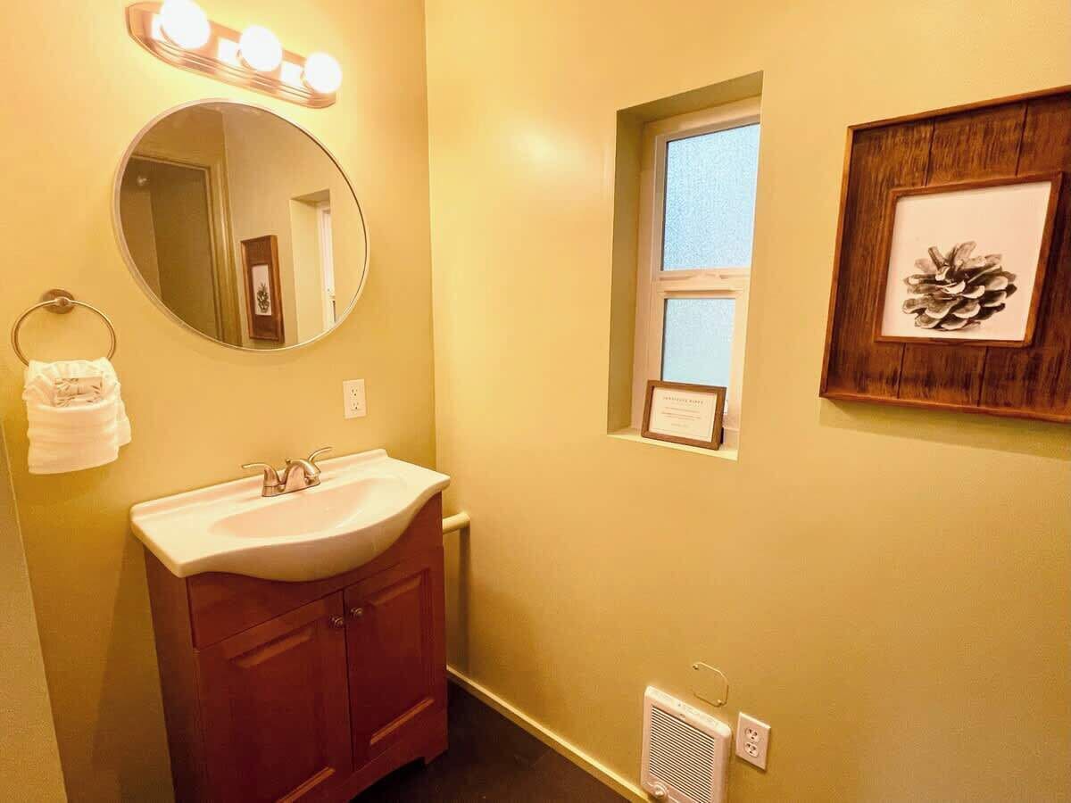 A bathroom with a vanity, a mirror with lights, a small window, and a framed pinecone decoration.