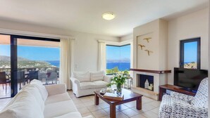 A spacious living room with a sofa, armchair, and a coffee table. There is a fireplace and a television. The room has large windows with a view of the sea.
