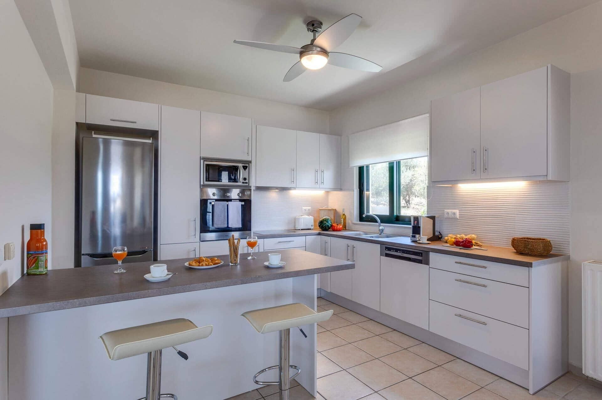 A modern kitchen with white cabinets, stainless steel appliances, a central island with bar stools, and a ceiling fan.