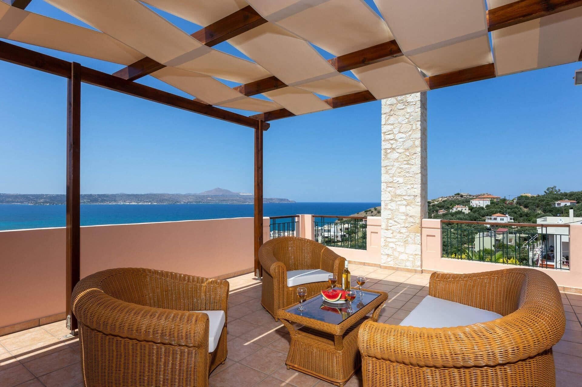 A balcony with wicker furniture, a table with a watermelon and wine glasses, and a view of the sea and distant hills.