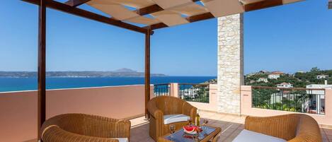 A balcony with wicker furniture, a table with a watermelon and wine glasses, and a view of the sea and distant hills.