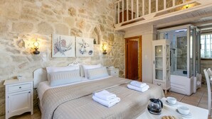A bedroom with a stone wall, a bed with white bedding, two bedside tables, a nightstand, a wooden door, and a glass shower enclosure. - Villa Minos by Villa Plus (Agia Pelagia)