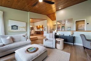 Una sala de estar moderna con un sofá, reposapiés y sillones, un comedor con una cocina al fondo, y obras de arte en las paredes.