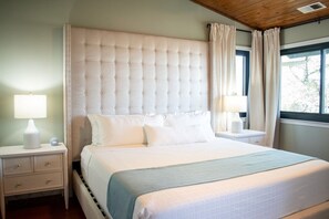 Une chambre à coucher avec une tête de lit capitonnée, un lit recouvert de draps blancs, deux tables de chevet avec des lampes et une fenêtre avec des rideaux.