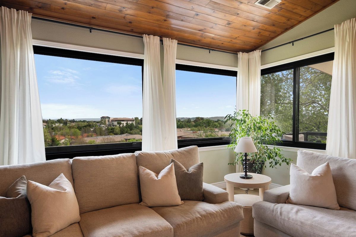 Habitación con sofá color beige, una mesita redonda con lámpara, y vistas a zonas verdes y edificios.