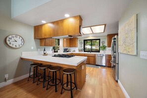 Una cocina moderna con una isla central, taburetes de bar, armarios de madera y electrodomésticos de acero inoxidable, además de un reloj de pared.