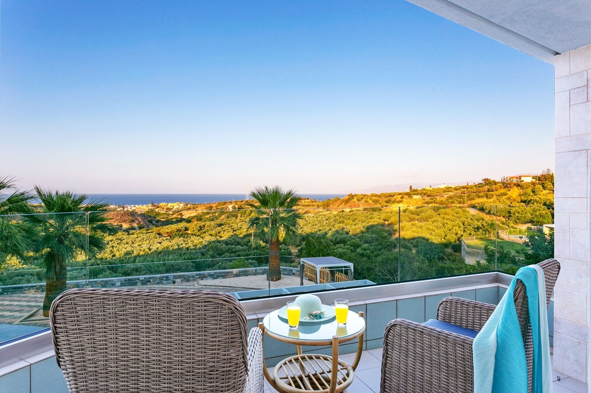 A balcony with wicker furniture, a table with drinks, and a view of the sea and greenery.