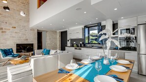 A modern kitchen with a dining area, featuring a wooden table set for dining, a large window with a view, and a stone wall. - Villa Potami Angela by Villa Plus (Apokoronos)