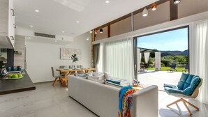 A modern living room with a sofa, a dining area, and a view of a garden and mountains. - Villa Potami Angela by Villa Plus (Apokoronos)