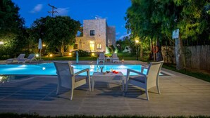 A swimming pool with lounge chairs and a table, surrounded by trees and a stone building with lights. - Villa Vincenzos by Villa Plus (Agia Pelagia)
