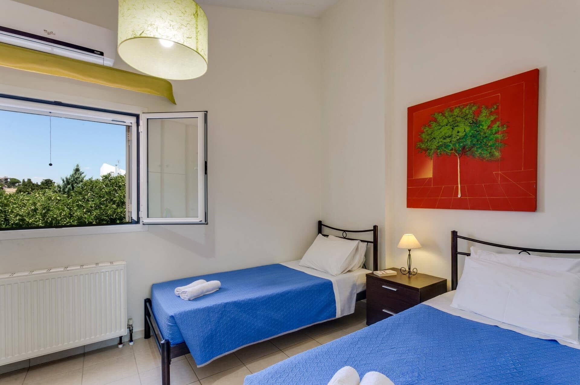 Une chambre d’hôtel avec deux lits, un radiateur, une fenêtre dont le volet est ouvert, une table de chevet avec une lampe et un tableau rouge représentant un arbre.