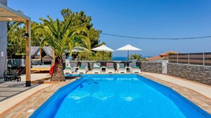 A swimming pool with lounge chairs and umbrellas, surrounded by palm trees and a stone wall. - Villa Omega by Villa Plus (Chersonisos)