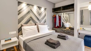 A modern bedroom with a herringbone-patterned headboard, a bedside table with a book, a bed with a grey quilt, and a mirror above a wooden dresser. - Villa Omega by Villa Plus (Chersonisos)