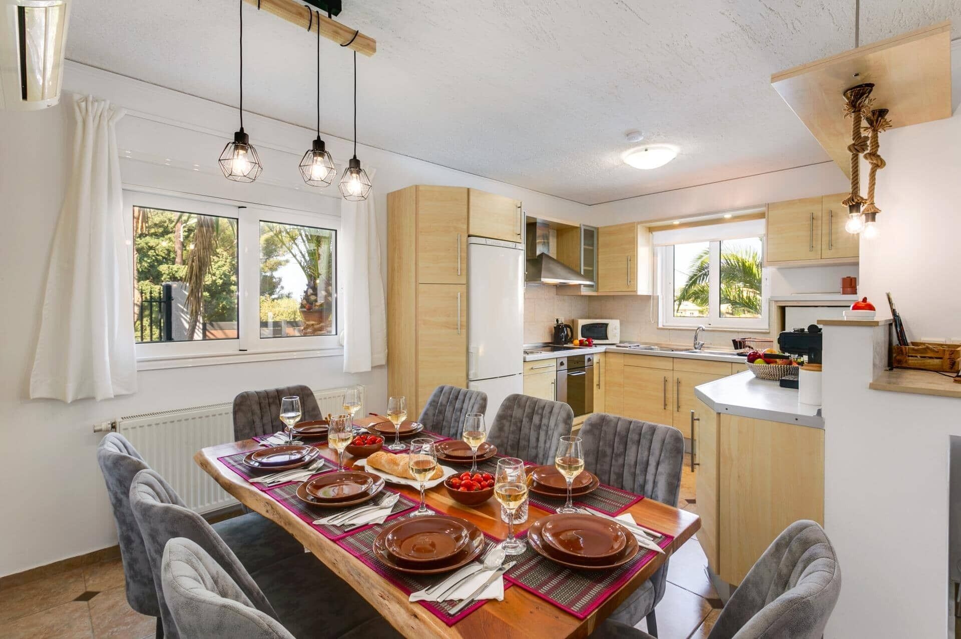 Un coin repas avec une table dressée pour six personnes, des meubles de cuisine, un réfrigérateur et une fenêtre donnant sur un paysage verdoyant.