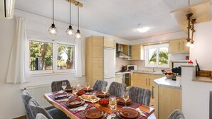 A dining area with a table set for six, kitchen cabinets, a refrigerator, and a window with a view of greenery. - Villa Omega by Villa Plus (Chersonisos)