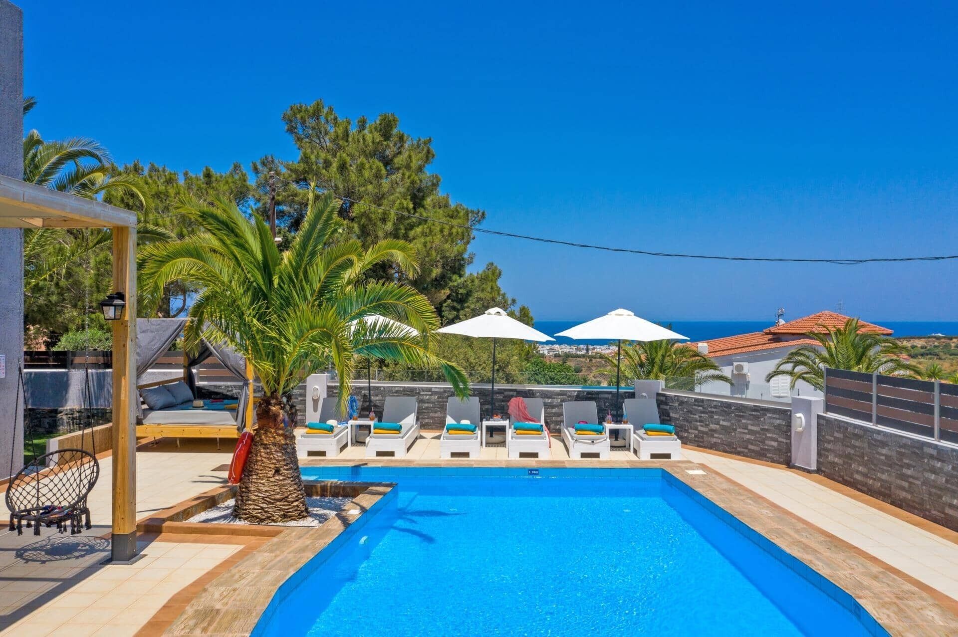 A pool area with lounge chairs and umbrellas, a palm tree, and a view of the sea.
