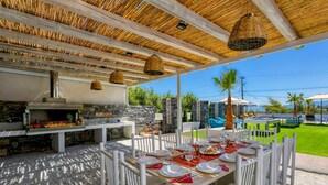 An outdoor dining area with a stone wall, a grill, and a pool in the background. - Villa Tropicana by Villa Plus (Hersonissos)