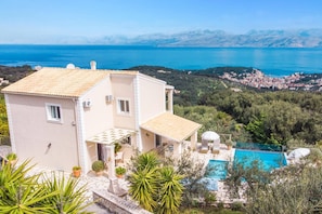Une maison à deux étages avec une piscine, entourée de verdure et offrant une vue sur une ville côtière.