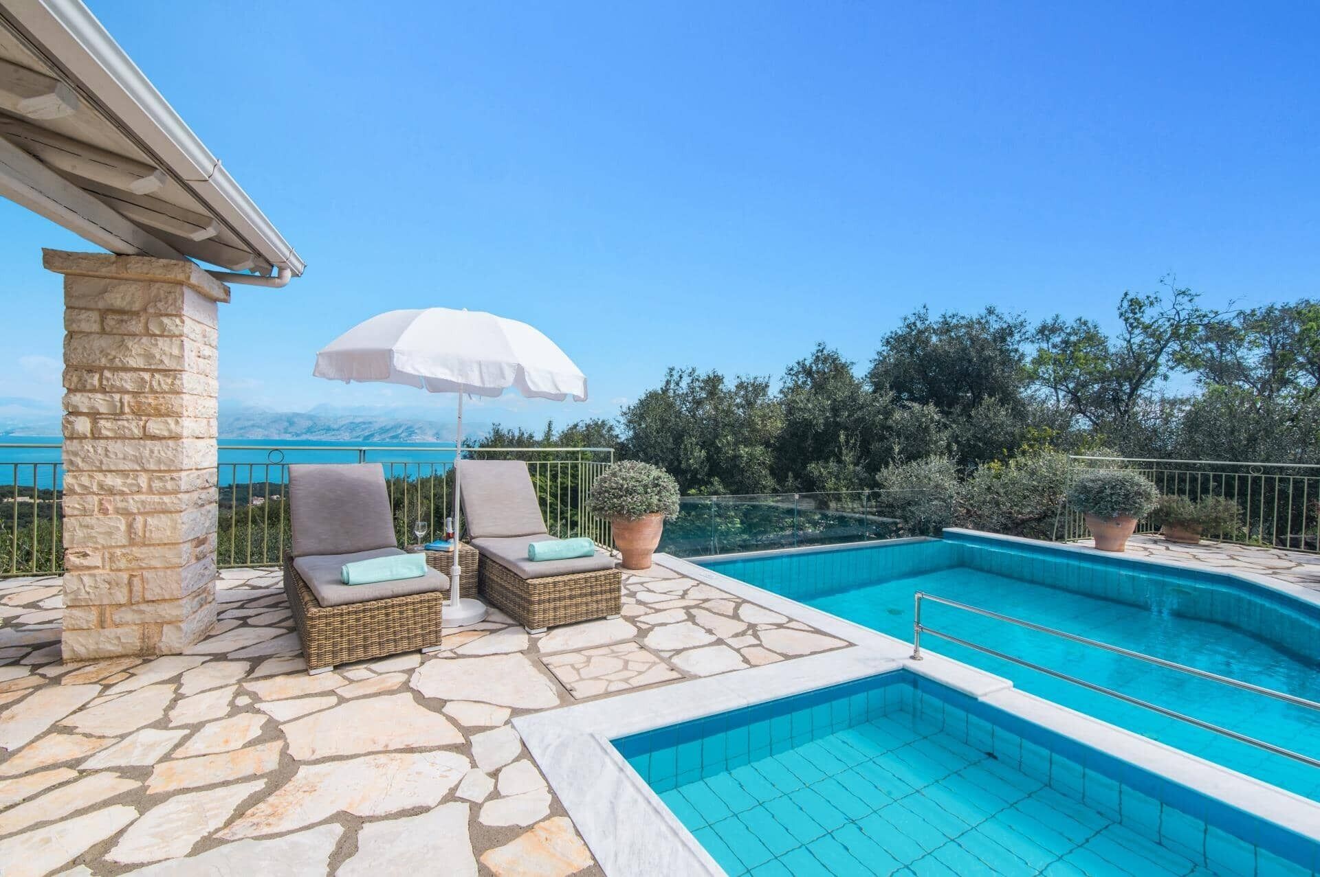 Ein Poolbereich mit Steinböden, zwei Korbstühlen, einem weißen Sonnenschirm und Blick auf das Meer und die Berge.