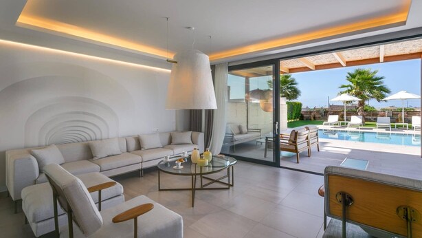 Een moderne woonkamer met een bank, salontafel en stoelen, een glazen deur die leidt naar een zwembadgedeelte met tuinmeubilair.