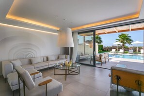 Un salon moderne comprenant un canapé, une table basse et des fauteuils, ainsi qu’une porte vitrée donnant sur un espace piscine aménagé avec du mobilier d’extérieur.