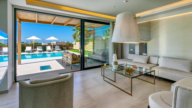 A modern living room with a glass coffee table, a sofa, and a view of a pool and outdoor seating area. - Villa Mariba White by Villa Plus (Malia)