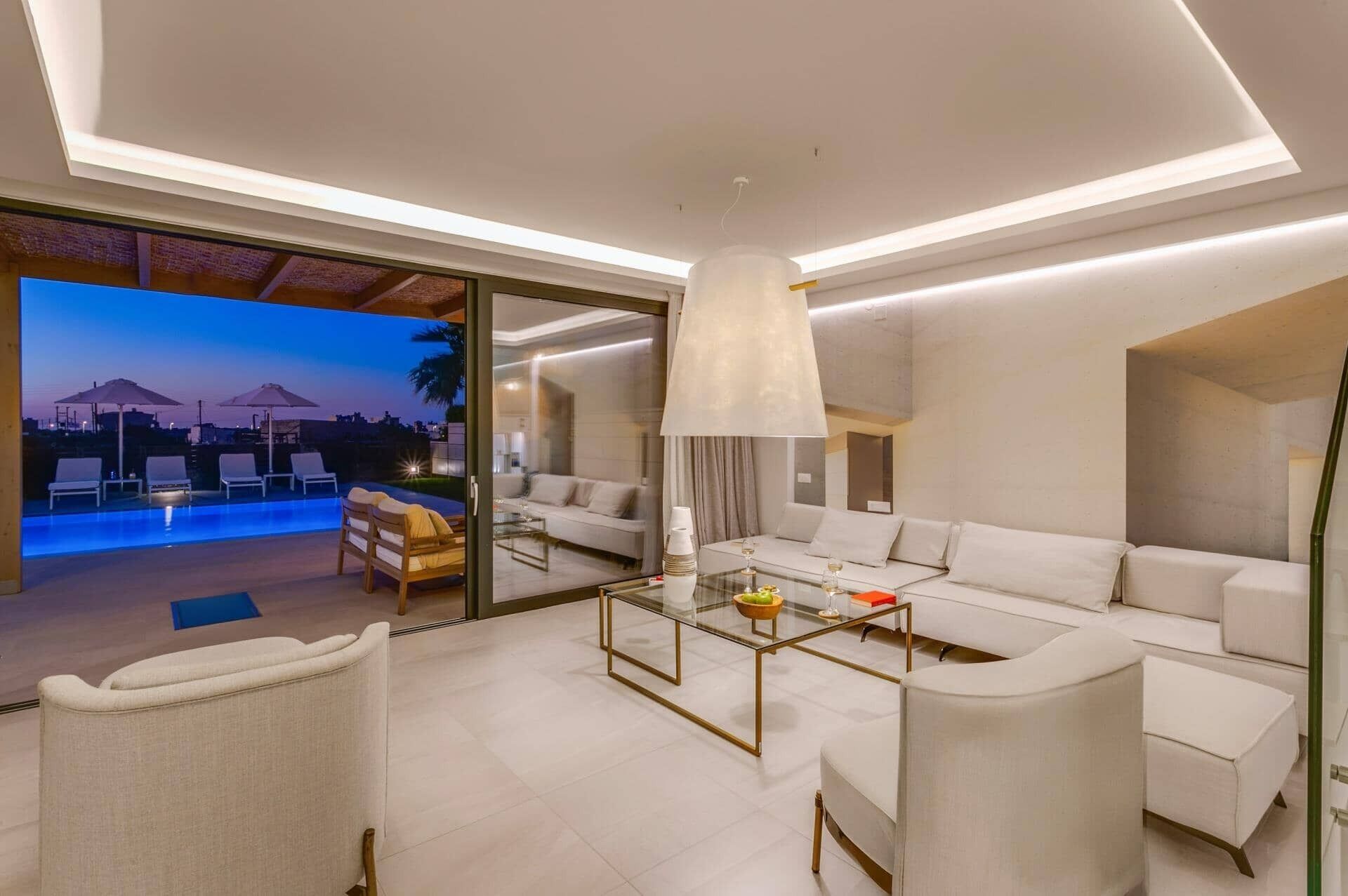 Una sala de estar moderna con una mesa de centro de cristal, sofás blancos y vista a una piscina al anochecer.
