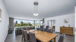 A modern dining area with a wooden table and leather chairs, large windows offering a view of a swimming pool and greenery, and a minimalist kitchen in the background. - Villa Casa Magnolia by Villa Plus (Vale Rebelho)