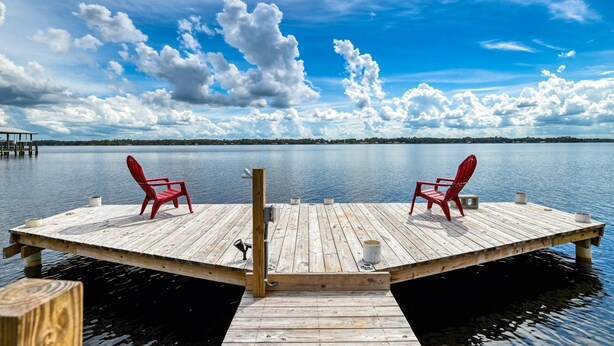 Un muelle de madera con dos sillas rojas mirando a un lago tranquilo bajo un cielo parcialmente nublado.