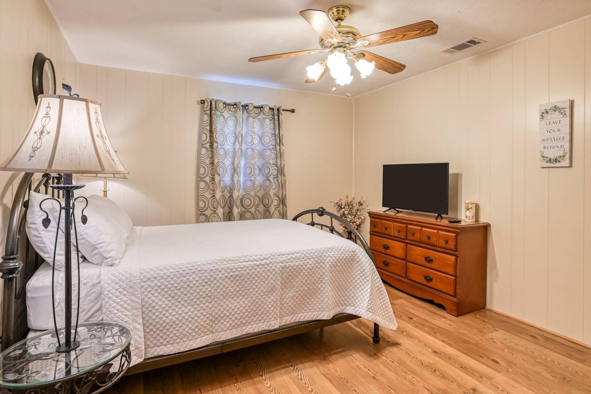 Une chambre à coucher comprenant un lit, une commode, un téléviseur, un ventilateur de plafond et une lampe.