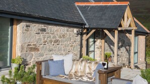 A stone building with a sloped roof, a wooden porch, and a seating area with cushions and a table.