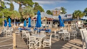 An outdoor dining area with tables and chairs, blue umbrellas, and a building with a red roof in the background.