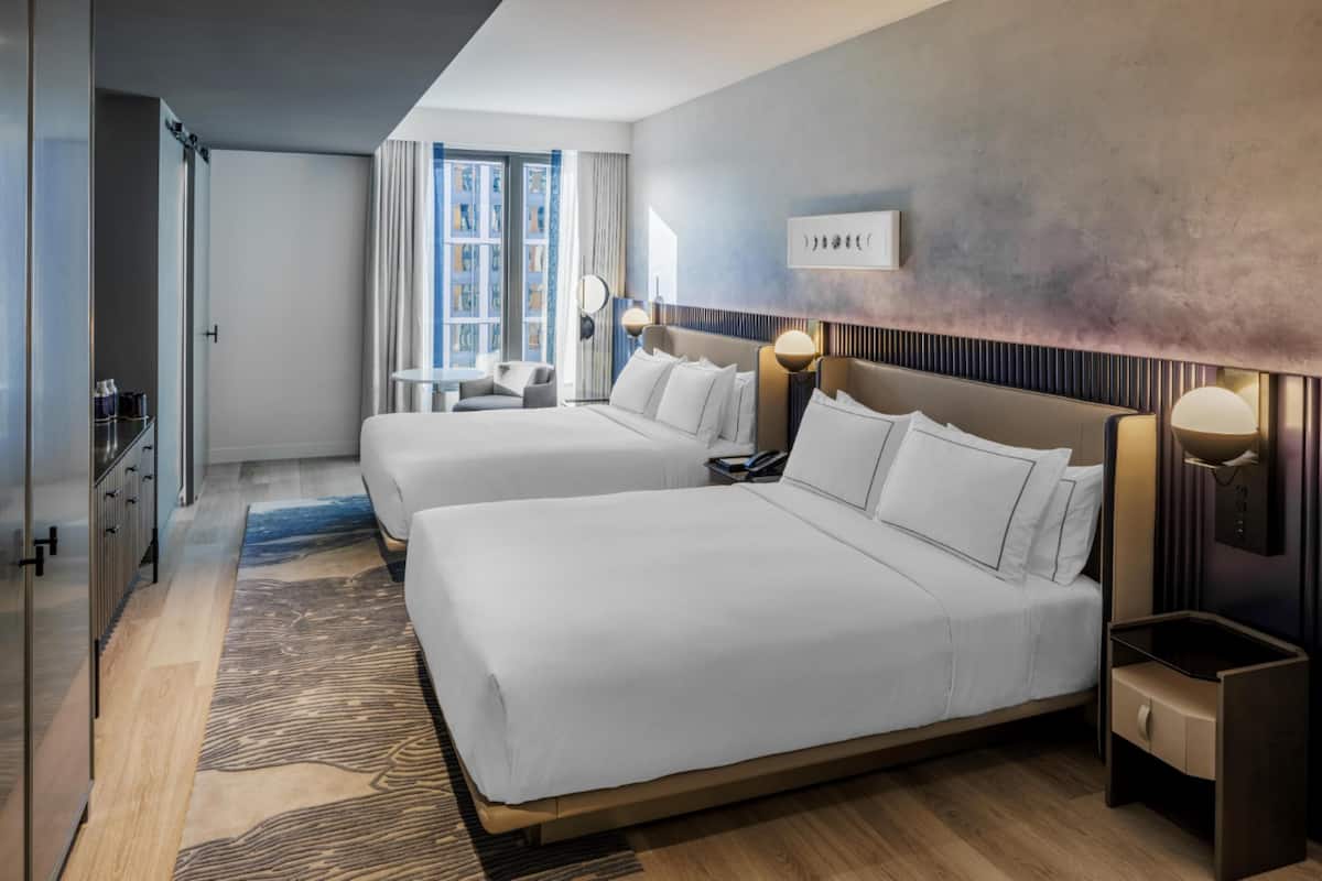 A hotel room with two beds, a desk, a chair, a lamp, and a window with curtains.