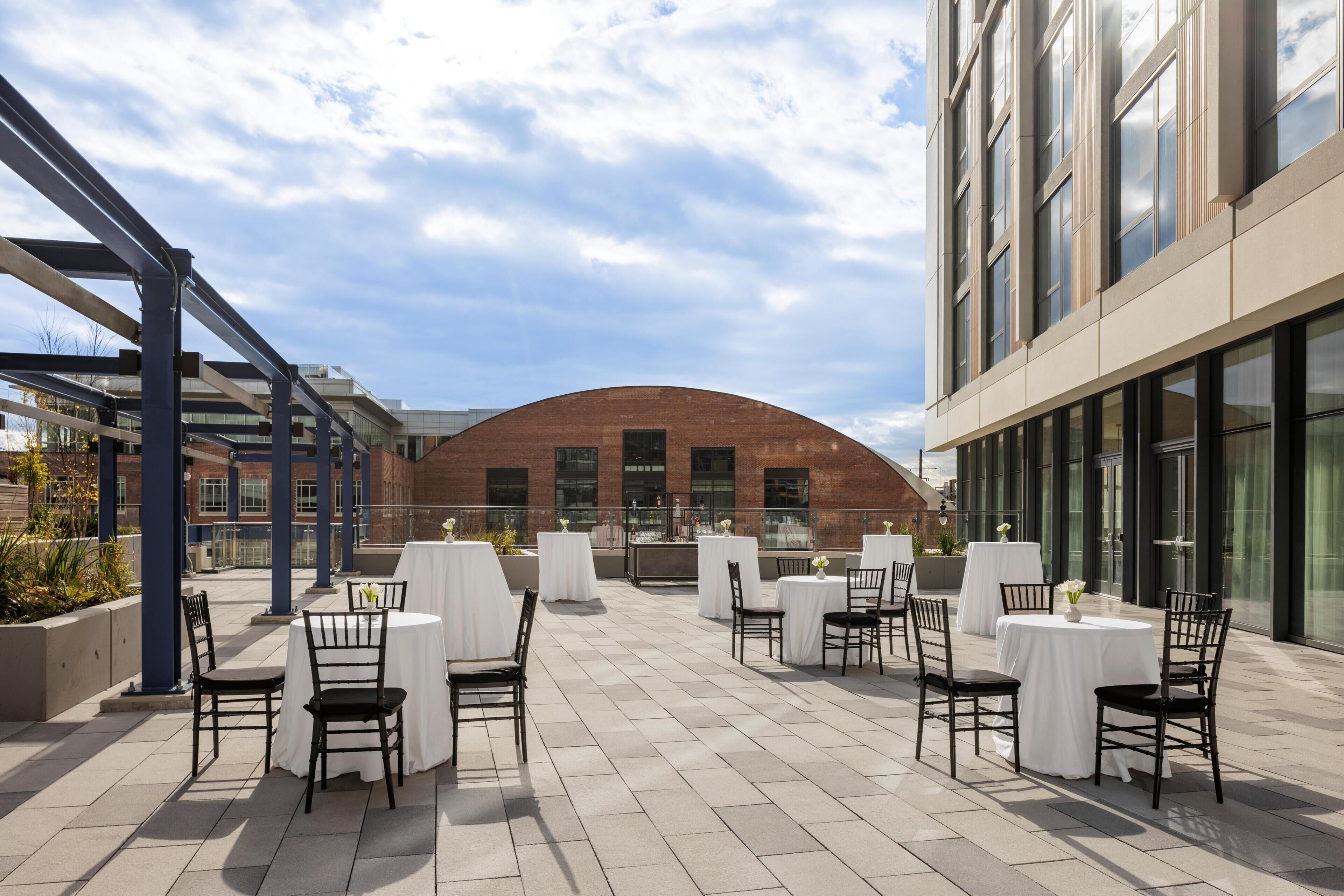Une terrasse extérieure avec des tables et des chaises, un bâtiment en briques en arrière-plan et un ciel dégagé.