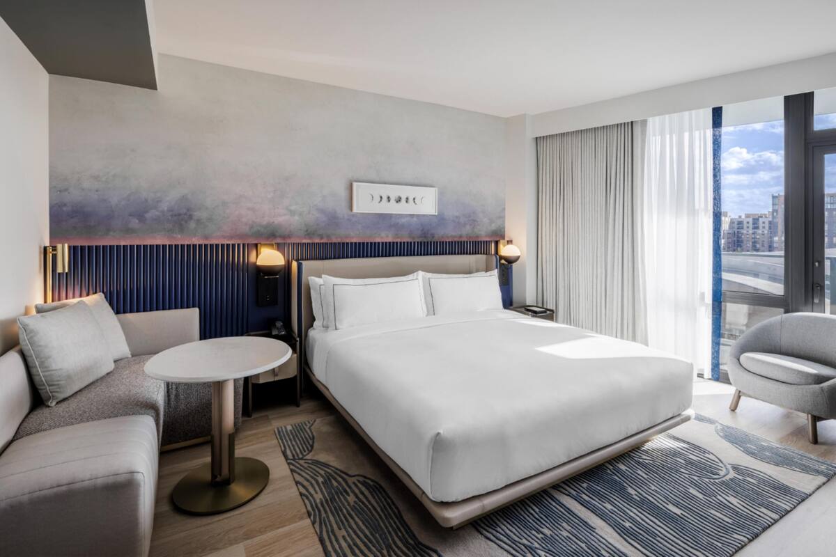 A modern hotel room with a large bed, a sofa, a small round table, and a view of the city through large windows.
