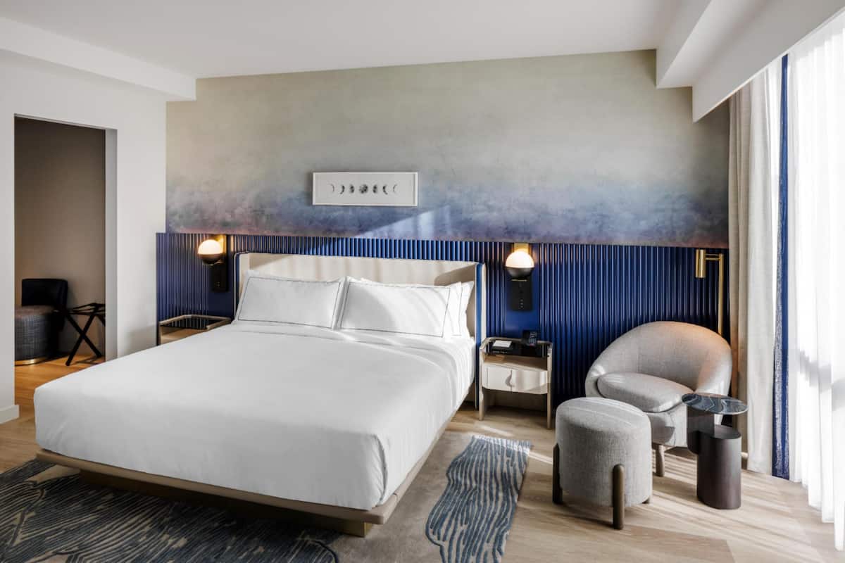 A modern hotel room with a large bed, a seating area with a chair and ottoman, a nightstand with a lamp, and a wall-mounted artwork.