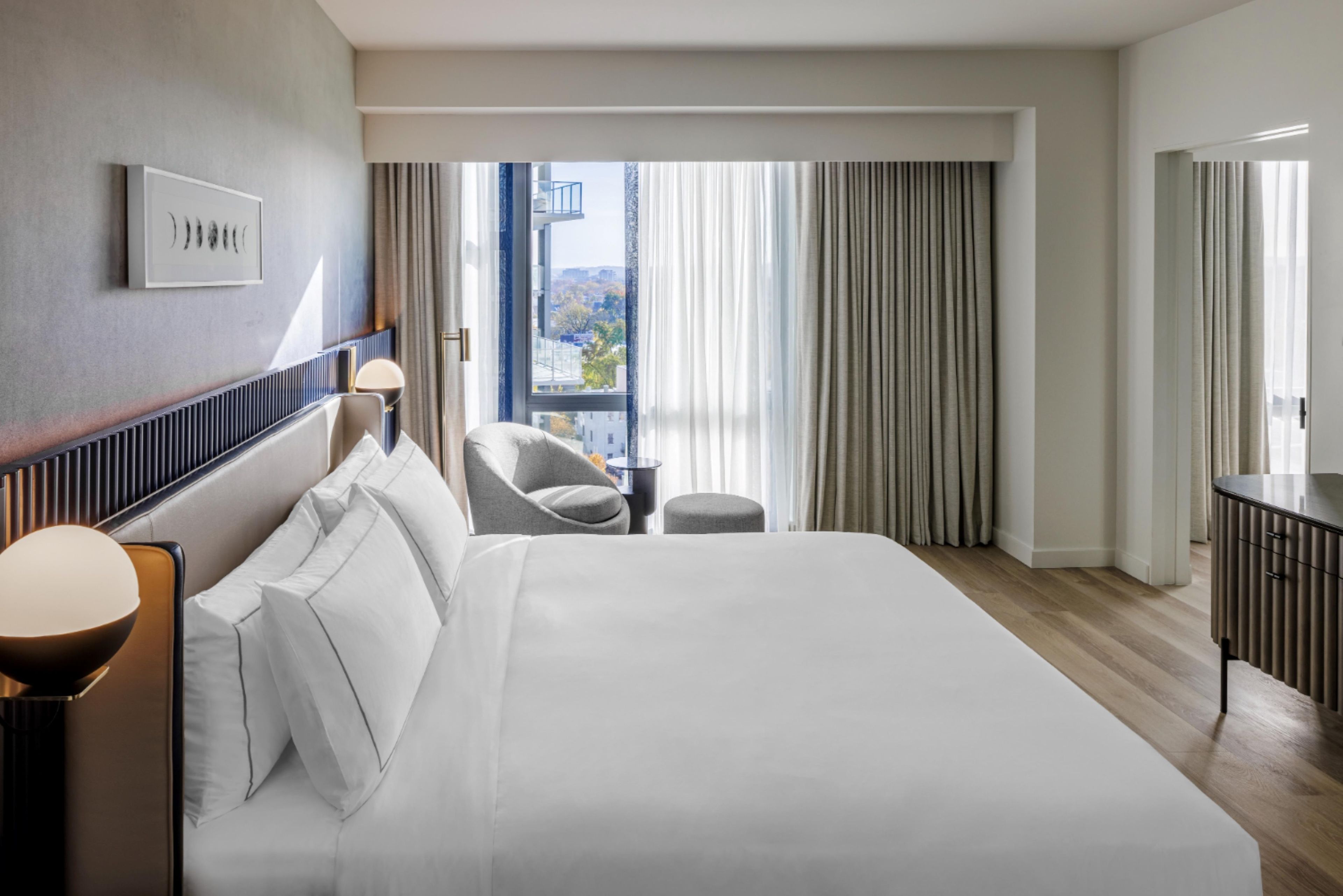 A hotel room with a large bed, a chair, a desk, and a view of the city through the window.