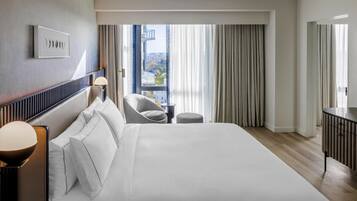 A hotel room with a large bed, a chair, a desk, and a view of the city through the window.
