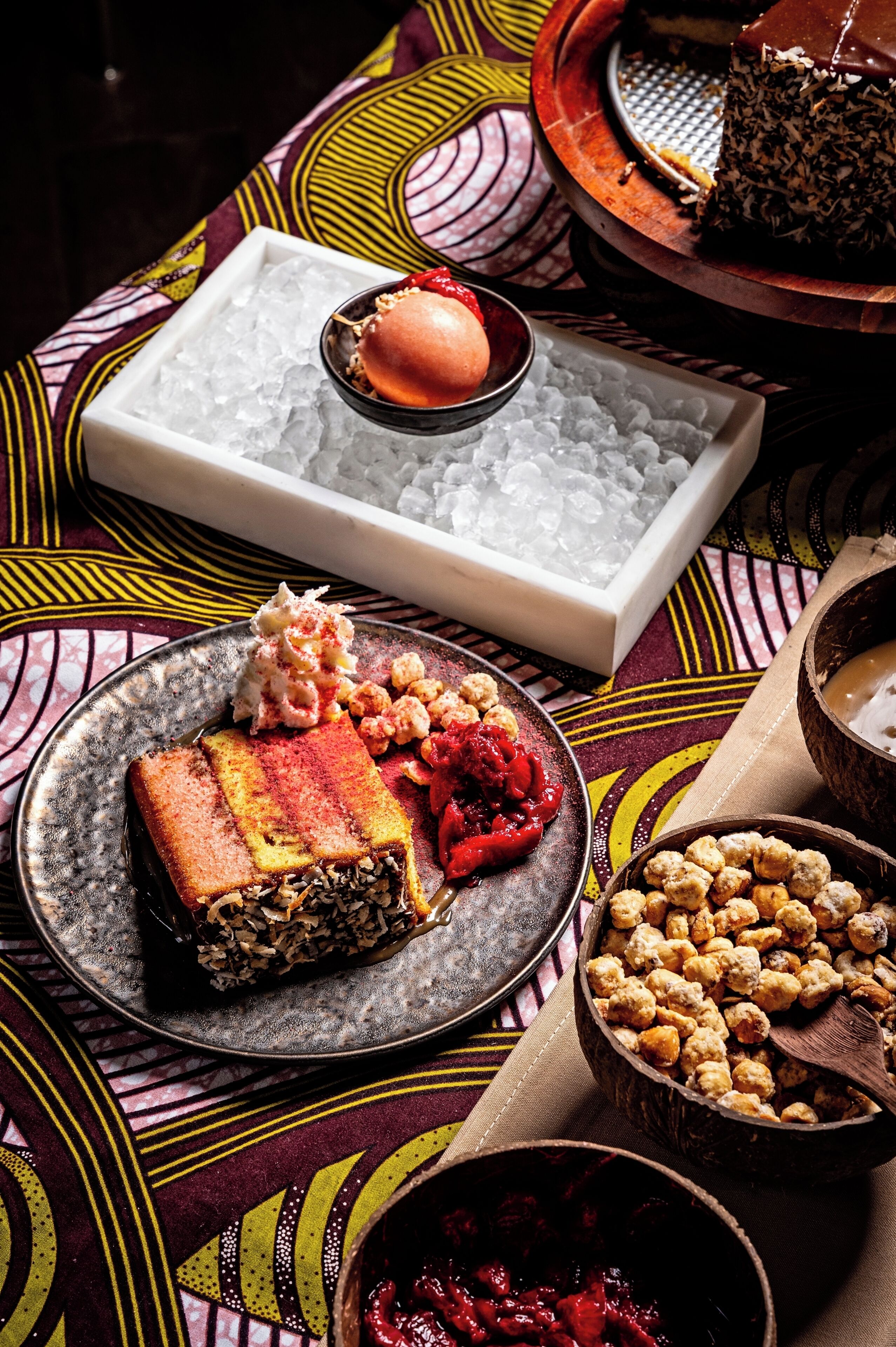 A dessert platter with various sweets and a bowl of chickpeas on a patterned tablecloth.