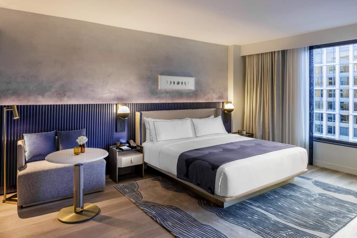 A modern hotel room with a bed, bedside table, lamp, and a view of the city.