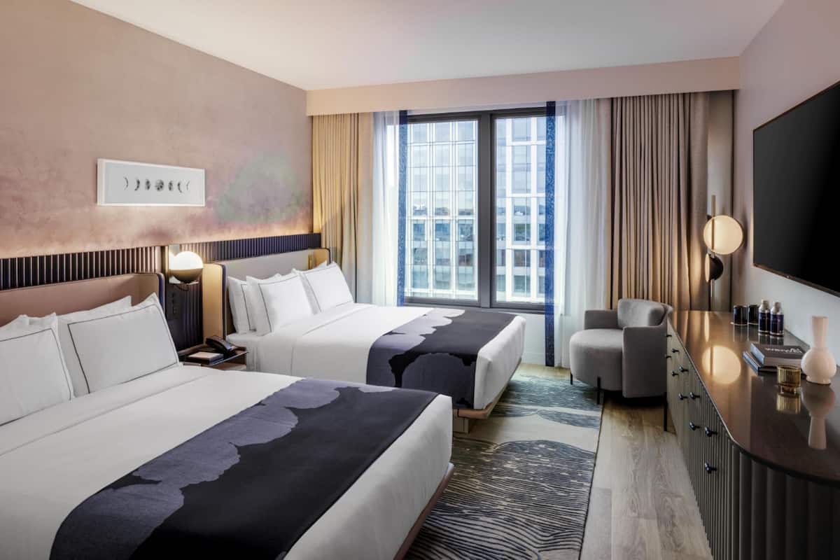 A hotel room with two beds, a desk, a chair, a TV, and a large window with curtains.