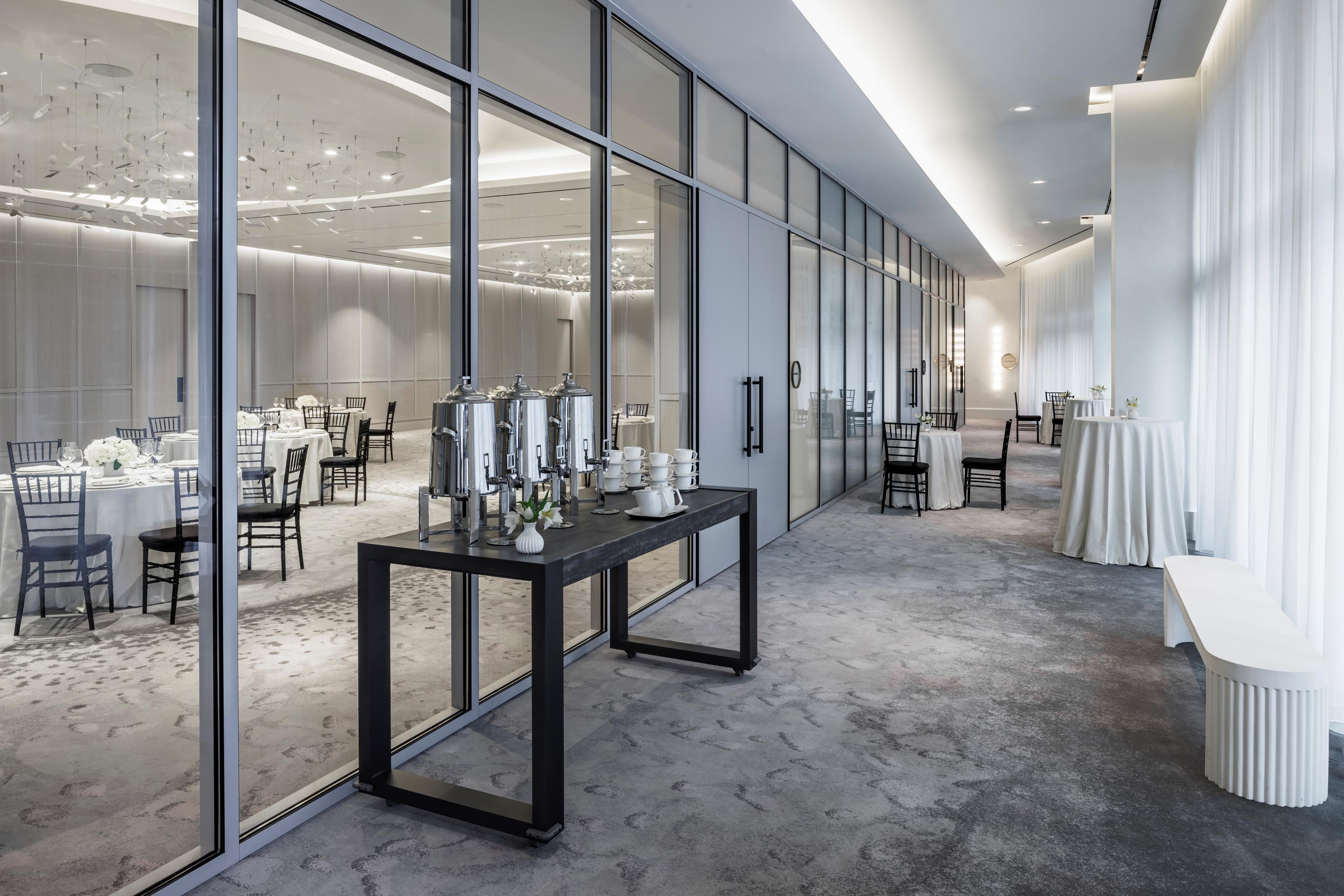 A modern hotel lobby with glass partitions, a central serving table, and multiple dining areas.