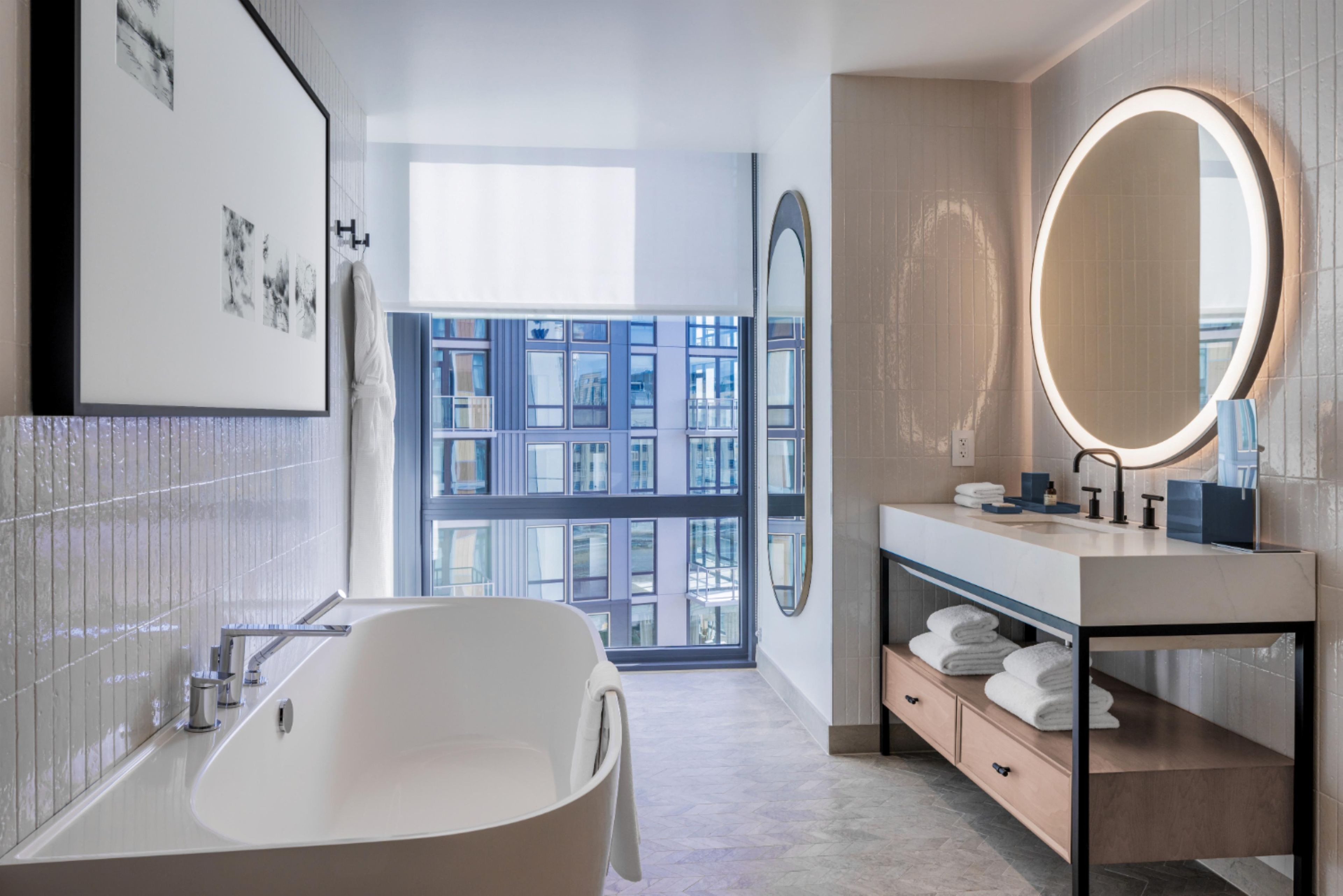 Un baño moderno con una bañera independiente, un espejo grande y vistas a la ciudad a través de un ventanal amplio.
