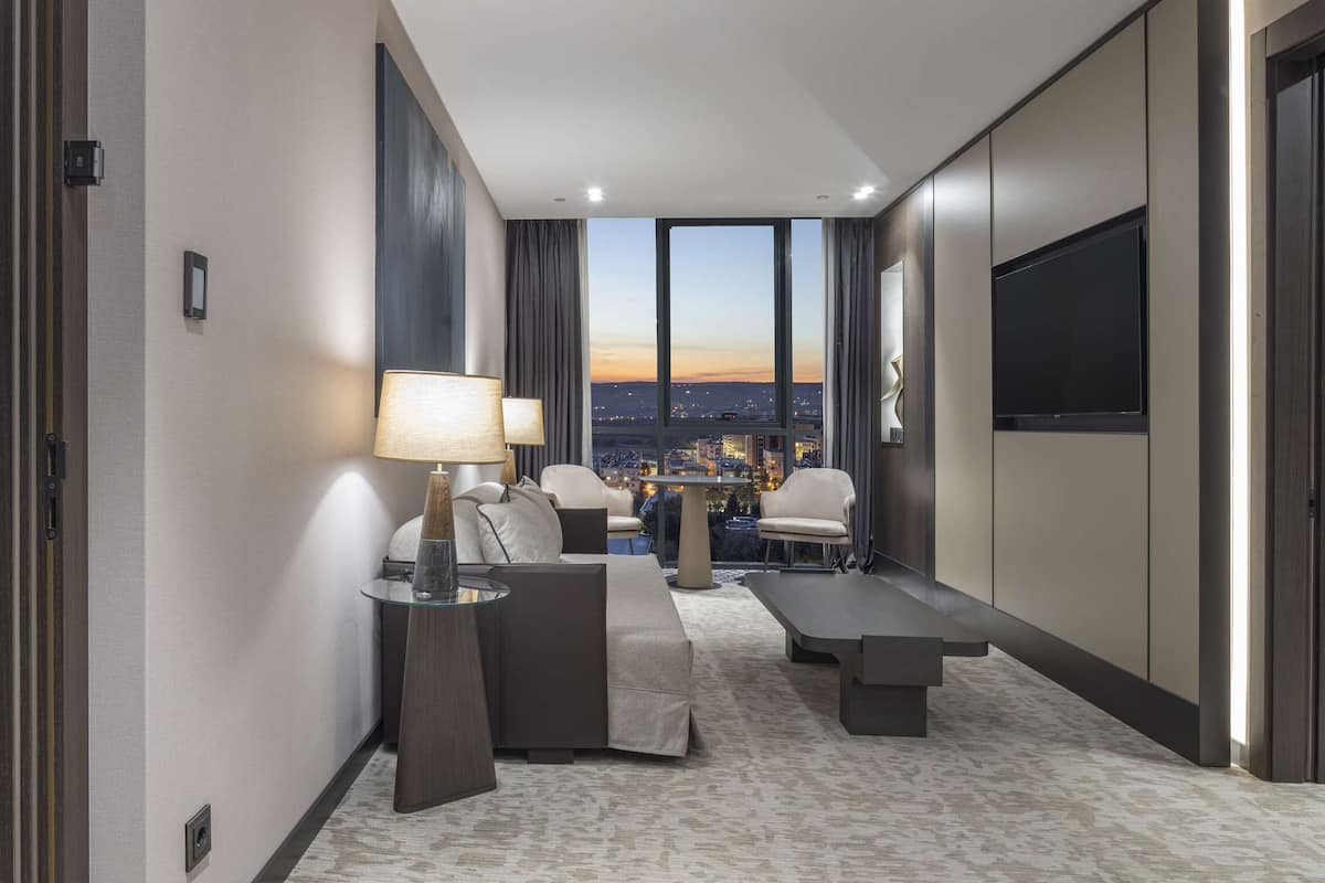 A modern hotel room with a bed, a seating area with a table and chairs, a large window with a city view, and a flat-screen TV.